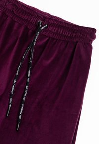 Bordeauxrode joggers gemaakt van geribbelde stof met een elastische tailleband. Voorzien van een trekkoord met zwarte merkdetails. De textuur lijkt zacht en comfortabel.