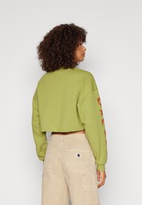 Cropped green sweatshirt with long sleeves, featuring red graphic prints on the left sleeve. Paired with beige cargo pants.