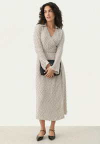 Beige Wickelkleid mit weißen Punkten, langen Ärmeln und V-Ausschnitt, kombiniert mit schwarzen spitzen Ballerinas und einer schwarzen Clutch.