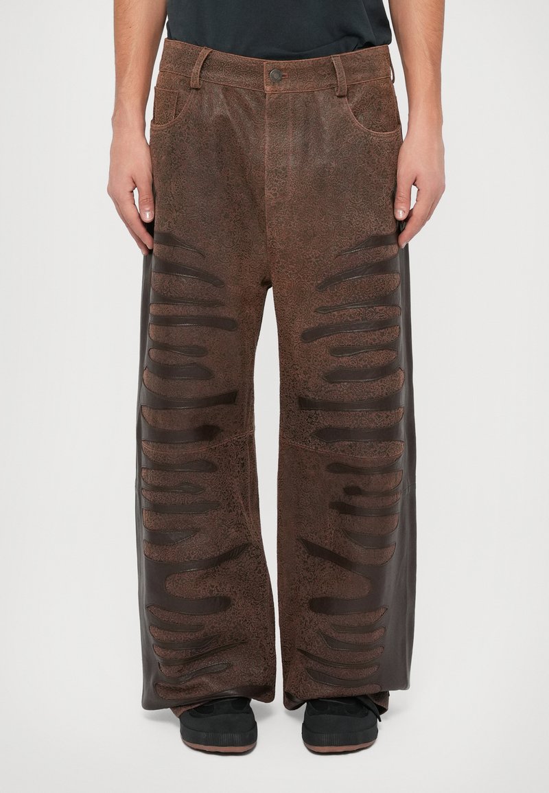 Brede bruine pantalons van textuurmateriaal, met een geprint oppervlak en zijuitsparingen in een donkere tint. Zwarte schoenen zichtbaar.