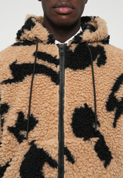 Close-up van een persoon die een tan en zwart gemêleerd fleece zip-up hoodie draagt met zwarte trekkoorden en een witte shirtkraag zichtbaar.