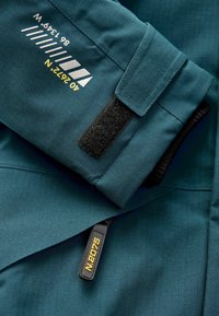 Türkise Stoffjacke mit einem Klettverschluss, strukturiertem Oberflächenmaterial und einem schwarzen Reißverschluss mit goldenen Details, versehen mit aufgedrucktem weißen und gelben Text.