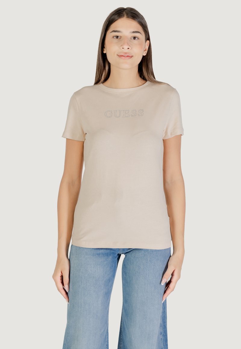 T-shirt beige a maniche corte con logo "GUESS". Tessuto morbido, collo a giro, silhouette aderente. Abbinato a jeans a gamba larga blu chiaro.