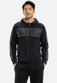 Homme portant un sweat à capuche noir zippé avec des détails de passepoil clairs et un pantalon noir assorti, les mains dans les poches, fond neutre.