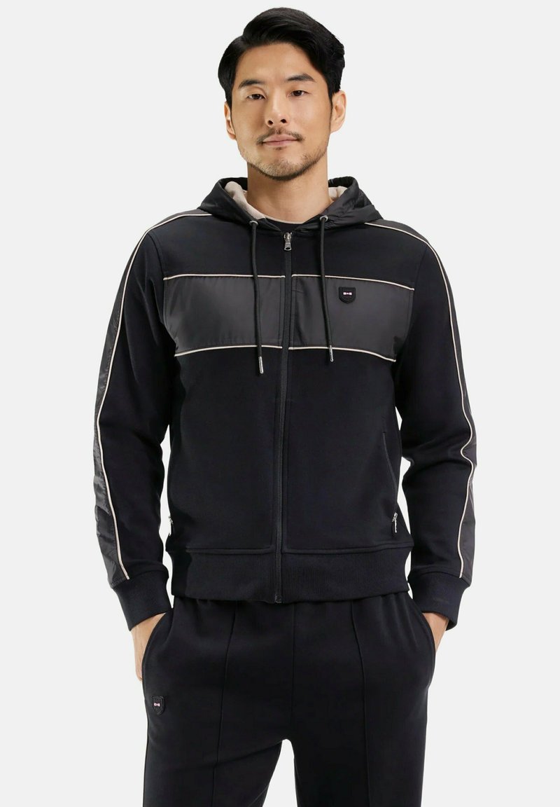 Homme portant un sweat à capuche noir zippé avec des détails de passepoil clairs et un pantalon noir assorti, les mains dans les poches, fond neutre.