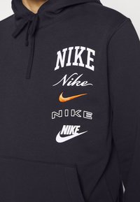 Svart huvtröja med flera Nike-logotyper i vit, orange och grå text. Designad med en framficka och dragsko på huva.