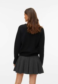 Kvinna med mellanmörkbrunt hår, klädd i en svart långärmad tröja och en mörkgrå plisserad kjol, vänd bort från kameran mot en vit bakgrund.