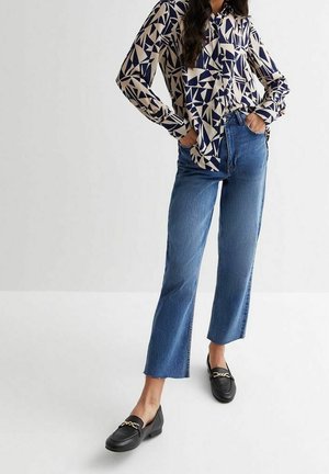 Kvinde iført blå bluse med geometrisk print, højtaljede cropped blå jeans og sorte loafers med guldkædedetalje, stående med hænderne i lommerne.