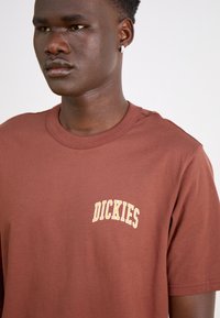 Brūns kokvilnas T-krekls ar īsām piedurknēm, apaļu kakla izgriezumu un dzelteno "DICKIES" logo izšūtu kreisajā krūšu daļā.