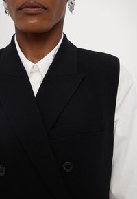 Gilet noir à double boutonnage avec des revers marqués, en tissu lisse, comportant une poche sur la poitrine et une chemise col blanc en dessous.