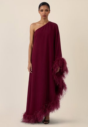 Kobieta w bordowej, sięgającej ziemi sukni z odkrytym jednym ramieniem, ozdobionej piórami wzdłuż dołu i rękawa, stojąca na jednolitym tle.