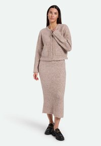Helles rosa, flauschiges Strickcardigan mit zwei vorderen Taschen, kombiniert mit einem passenden langen Rock. Beide Teile weisen eine weiche, strukturierte Oberfläche auf. Schwarze Slipper.