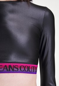 Černý dlouhý rukáv cropped top vyrobený z lesklé, hladké látky s odvážným, barevným elastickým páskem, na kterém je vytištěno "JEANS COUTURE" černým písmem.