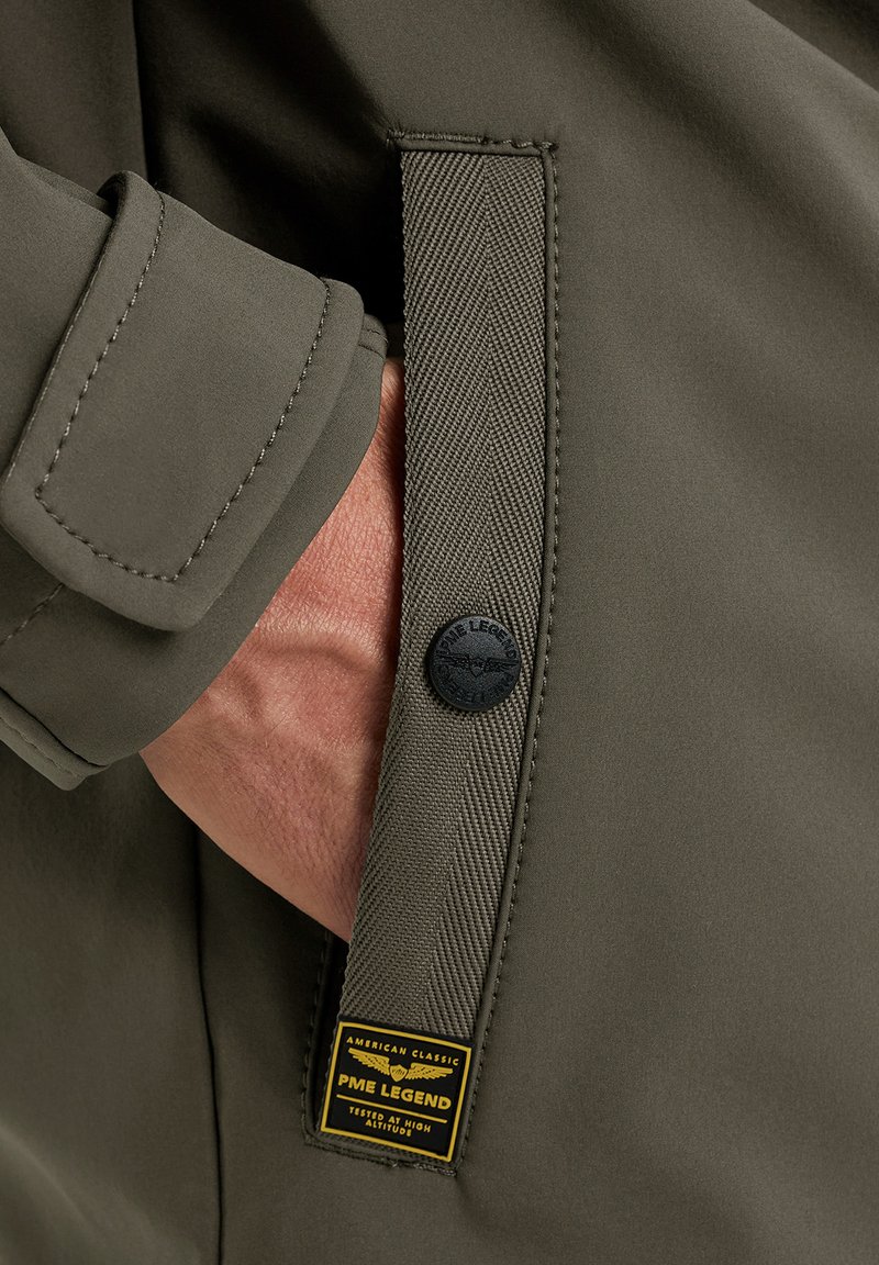 Main dans la poche d'une veste vert olive avec une sangle en tissu, un bouton noir, et une étiquette jaune PME Legend indiquant "American Classic Tested at High Altitude."