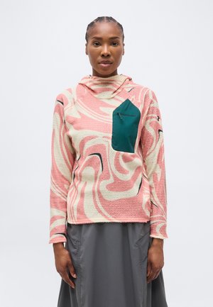 Femme portant une polaire rose et crème à motifs avec une poche poitrine vert foncé et une jupe grise, debout devant un fond blanc uni.