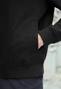Juodas flisas su užtrauktuku, su raukšlėtomis rankovėmis ir šone kišene. Minkšta tekstūra ir prigludusi siluetas.