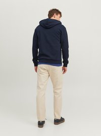 Navy hettegenser med kangaroo-lomme foran, beige bukser og svarte sneakers. Teksturen ser myk ut; hettegenseren har snøring og stor hette.