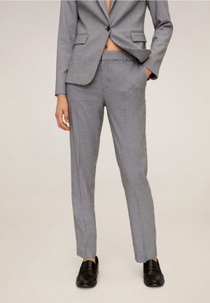 Kobieta ubrana w szare spodnie garniturowe w kratę i pasującą marynarkę, stojąca z jedną ręką w kieszeni w czarnych mokasynach.