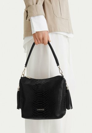 Dłoń trzymająca czarną teksturowaną torebkę z frędzlami, ubrana w beżową kurtkę i białe spodnie na jednolitym tle.