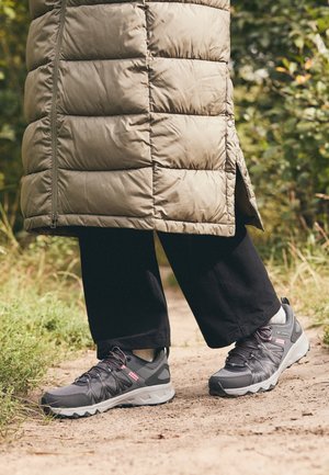 Personne portant un pantalon noir, des chaussures de randonnée grises et un manteau matelassé vert marchant sur un sentier de terre entouré de verdure.