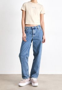 Persona che indossa una t-shirt beige corta di Calvin Klein, jeans blu larghi e sneakers con plateau rosa chiaro, in piedi su un pavimento semplice.