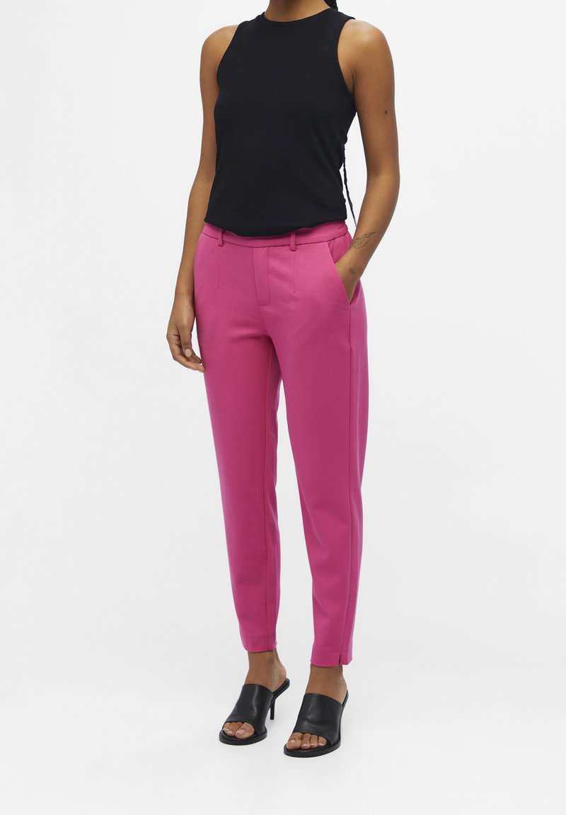 Femme portant un haut noir sans manches, un pantalon rose vif ajusté et des mules à talons ouvertes noires, debout, une main dans la poche.
