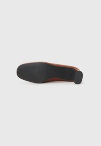 Chaussure habillée en cuir marron retournée pour montrer la semelle noire texturée et le talon rectangulaire sur un fond clair uni.