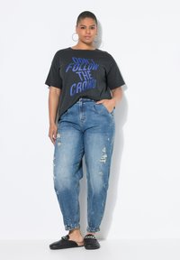 Camiseta negra gráfica con texto azul; pantalones vaqueros sueltos azul claro con efecto desgastado; zapatos de slide negro con acento de cadena; grandes pendientes de aro.