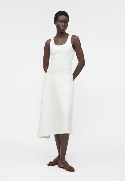 Γυναίκα με σκουρόχρωμο δέρμα φοράει λευκό αμάνικο τοπ και ασύμμετρη λευκή μίντι φούστα, στέκεται με τα χέρια πίσω από την πλάτη φορώντας καφέ σανδάλια.