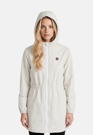 Femme aux longs cheveux blonds portant une veste à capuche beige clair avec une fermeture éclair frontale et un petit logo rond rouge et bleu sur la poitrine.