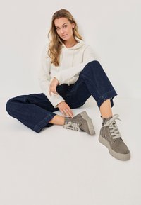 Graue Wildleder-Stiefeletten mit dicken, strukturierten Sohlen und hellgrauen Schnürsenkeln. Das Model trägt einen cremefarbenen Hoodie und dunkelgraue, weite Jeans und sitzt auf einer weißen Fläche.
