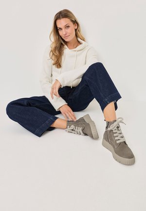 Bottines en daim gris avec des semelles épaisses et texturées, et des lacets gris clair. Le modèle porte un sweat à capuche crème et un jean large noir, assis sur une surface blanche.