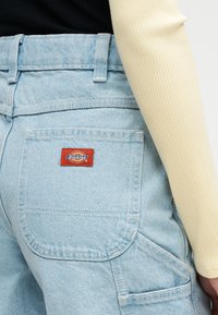 Ανοιχτό μπλε παντελόνι τζην με πίσω τσέπη που φέρει ετικέτα "Dickies", φορεμένο με κρεμ ριμπ μπλούζα με μακριά μανίκια και μαύρο τοπ.