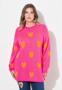 Różowy sweter z dzianiny z pomarańczowymi wzorami w kszta�łcie serc, okrągły dekolt, długie rękawy i ściągacze na końcach rękawów. Połączony z niebieskimi dżinsami z akcentem w kształcie serca.