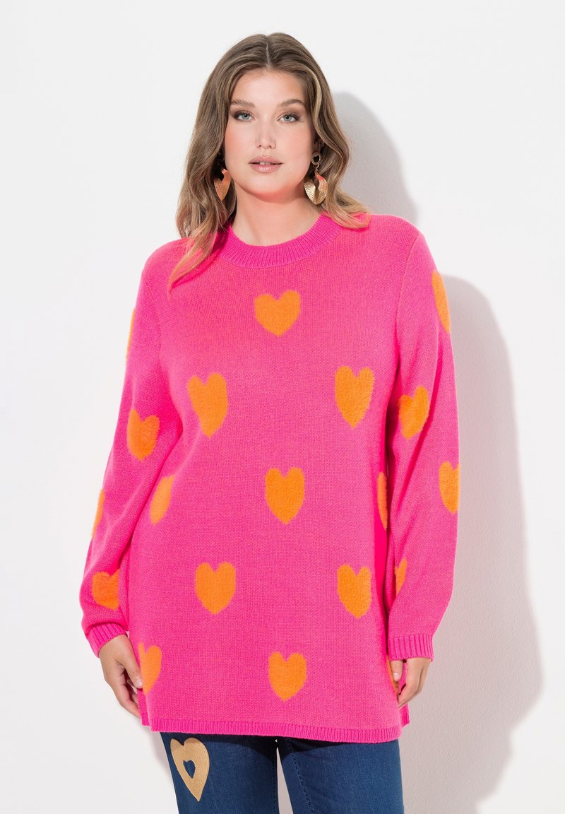 Różowy sweter z dzianiny z pomarańczowymi wzorami w kształcie serc, okrągły dekolt, długie rękawy i ściągacze na końcach rękawów. Połączony z niebieskimi dżinsami z akcentem w kształcie serca.