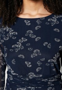 Marineblaues Kleid mit weißem Blumenmuster, aus strukturiertem Stoff und mit geraffter Taillenpartie. Rundhalsausschnitt und lange Ärmel sind sichtbar.