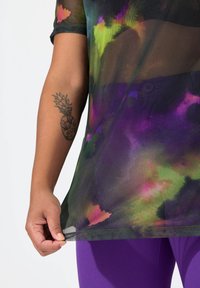 Gennemsigtig, multifarvet top med livlige grønne, lilla og pink mønstre. Korte ærmer og rundet kant, parret med lilla leggings.