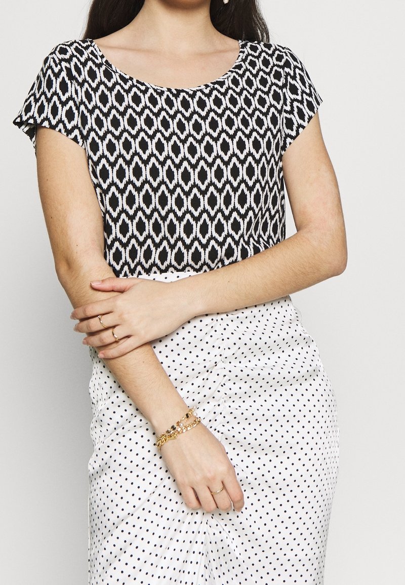 Haut à manches courtes à motif noir et blanc avec un col rond, associé à une jupe blanche à pois et des bracelets dorés au poignet.