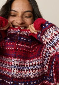 Rode gebreide trui met ingewikkelde meerkleurige patronen, geribbelde manchetten en een hoge kraag. De textuur oogt warm en gezellig.