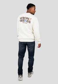 Sudadera de cuello redondo en color crema con el logotipo "WRONG FRIENDS" en la parte posterior en colores contrastantes. Combinada con jeans oscuros y zapatillas deportivas.