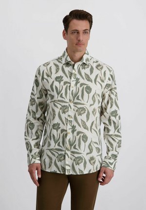 Mann trägt ein weißes Hemd mit grünen Blumenmustern und braune Hose, stehend vor einem einfachen hellen Hintergrund.