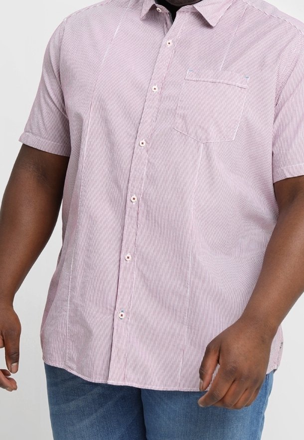 Homme portant une chemise à manches courtes, violette claire, à boutons et fines rayures verticales, avec un jean bleu, debout les bras détendus.