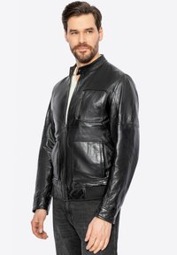 Chaqueta de cuero negra con cuello alto, cierre frontal de cremallera, múltiples bolsillos y una textura suave. Presenta puños ajustados y un diseño elegante.