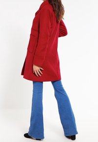 Abrigo de mezcla de lana roja con cuello de muesca, bolsillos laterales y mangas largas, combinado con vaqueros acampanados azules y calzado negro.