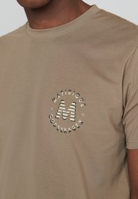 Bežinis medvilninis marškinėlis su apvaliu logotipu, kuriame yra "MATINIQUE COPENHAGEN" ir dryžuotas "M" dizainas, trumpomis rankovėmis ir klasikinio kirpimo.