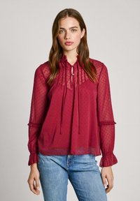 Blusa trasparente borgogna con texture a pois, scollatura arricciata e polsini. Maniche lunghe con dettaglio a bottoni. Abbinata a jeans blu.