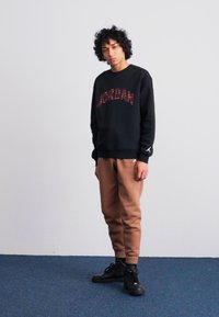 Felpa nera con la scritta "JORDAN" in caratteri rossi, abbinata a pantaloni jogger marrone chiaro. Stilizzata con sneaker alte nere. Texture liscia.