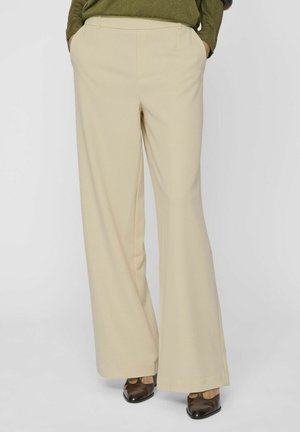 Beige wijde broek van een gladde stof, met zijzakken en een op maat gemaakte tailleband. Gepaard met donkerbruine schoenen.