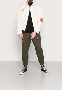 Persoon draagt een witte jas met een oranje bloem en smiley face patches, een zwart shirt, groene cargobroek en zwarte hoge sneakers.
