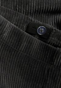 Zwart corduroy stof met textuur en verticale ribbels. Beschikt over een prominente knoopsluiting met een transparante afwerking en een elastische band.